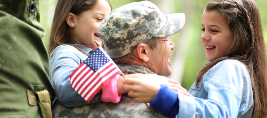 (unoptimized) Family welcomes home a USA army soldier. The children excitedly hug father holding American flags.