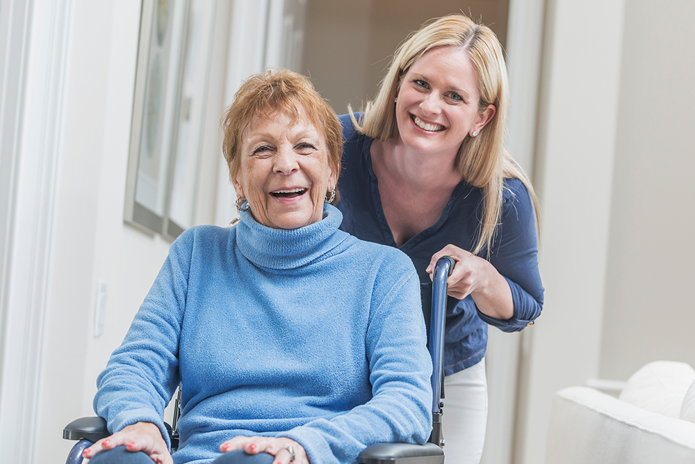 (unoptimized) elderly lady in wheelchair wearing blue long sleeve turtleneck sweater with contentedly happy smile (possibly laughing) with younger lady leaning into the picture wearing darker blue V-neck shirt smiling for the picture.