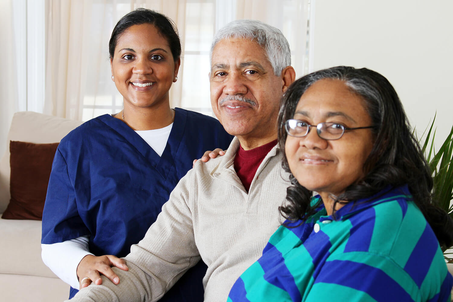 Older man in grey sweater with maroon under shirt sits next to lady in blue and green stiped shirt with glasses while nurse in dark blue scrubs stands behind man's shoulder (all smiling)