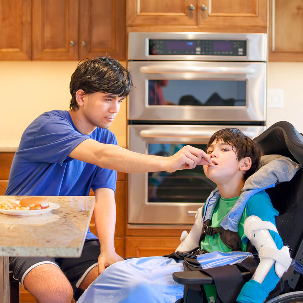 brother in blue shirt feeding weelchair bound younger brother in green and blue shirt in contemporary kitchen with double oven in the background, wood cabinets and granite countertop