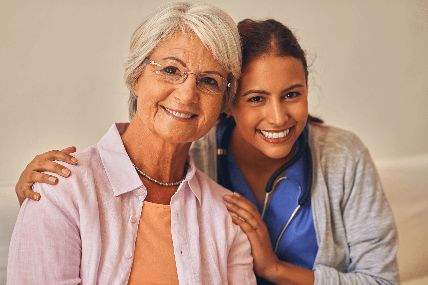 elderly lady wearing orange blouse with white rough silk over shirt, short grey hair parted over right eye with glasses and inquisitive smile leaning into a hug by a younger nurse with broad smile blue scrubs, stethiscope and grey cotton over shirt