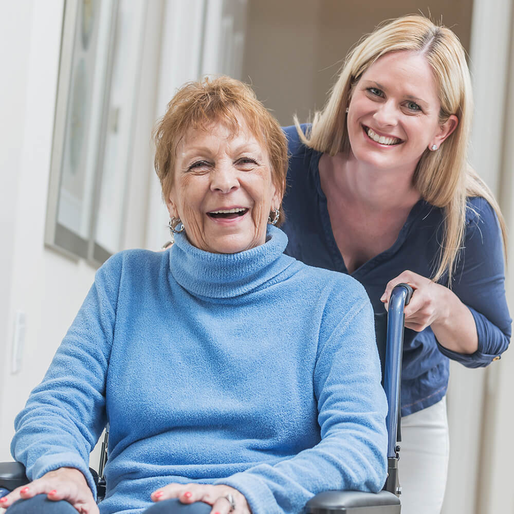 elderly lady in wheelchair wearing blue long sleeve turtleneck sweater with contentedly happy smile (possibly laughing) with younger lady leaning into the picture wearing darker blue V-neck shirt smiling for the picture.